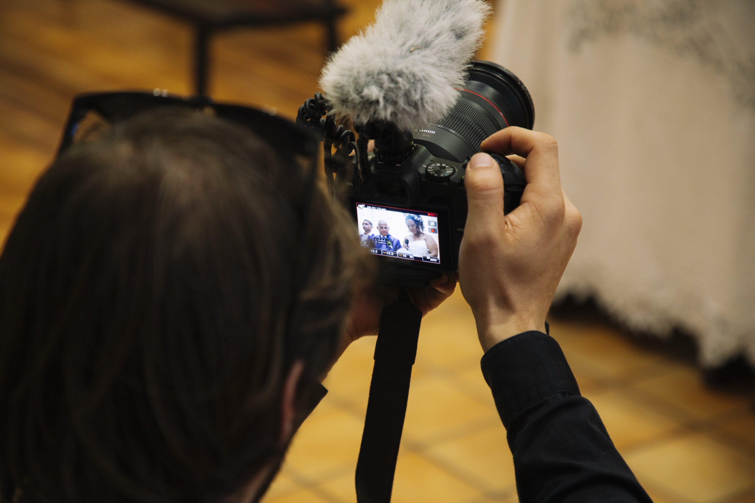 Une personne est vue de dos, tenant une caméra noire avec un micro à poils gris monté dessus, en train de filmer ou prendre des photos