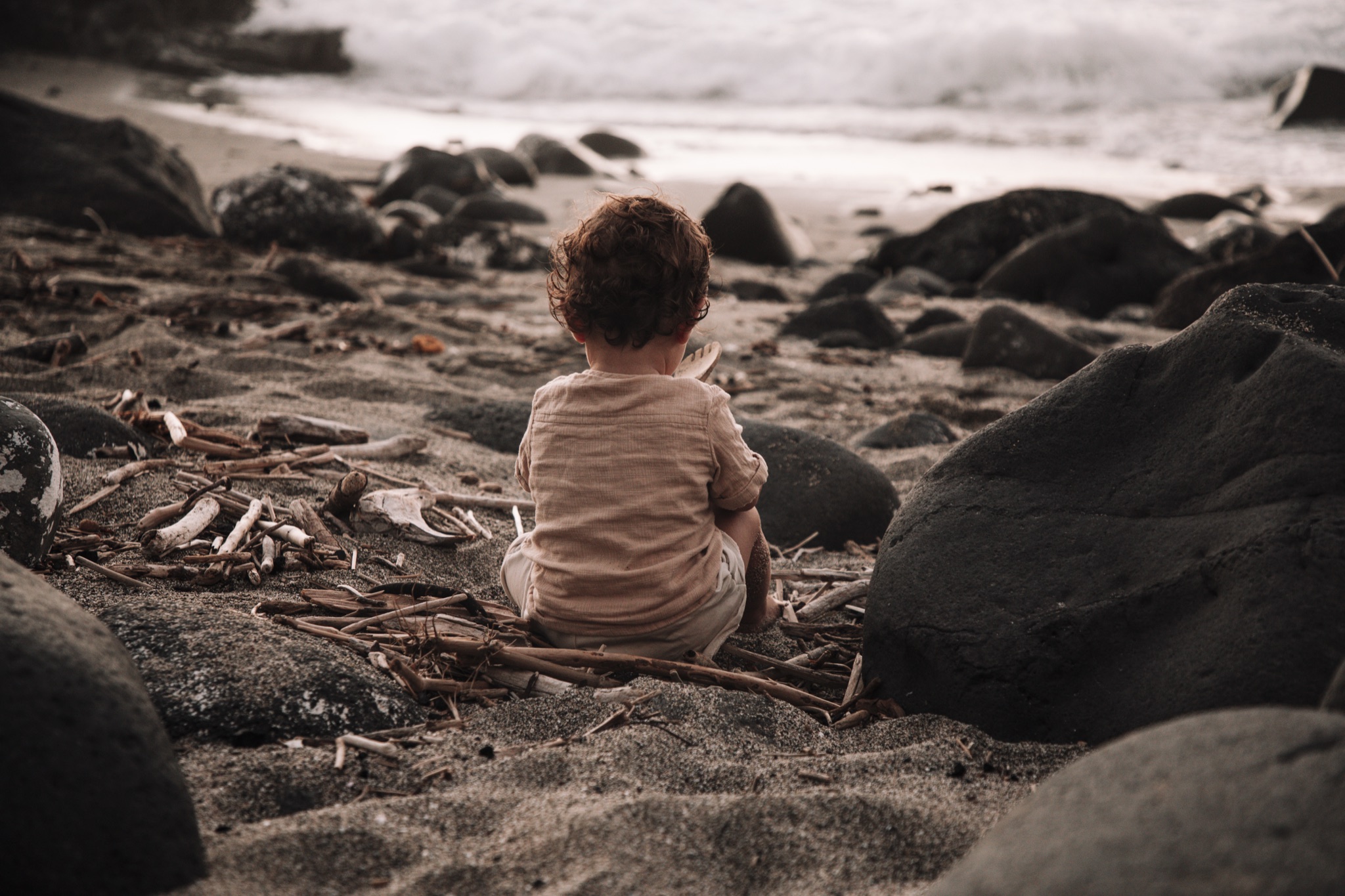 Un jeune enfant aux cheveux bouclés est assis dos à la caméra sur une plage rocailleuse, entouré de bois flottés et de vagues qui s'éloignent dans le