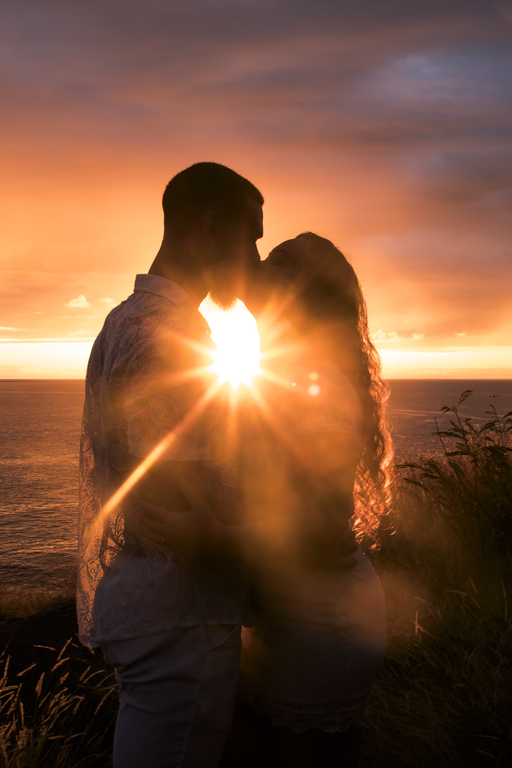 Deux silhouettes humaines se tiennent face à face au bord d’un paysage côtier, leurs corps enveloppés par une lumière dorée intense émanant du soleil