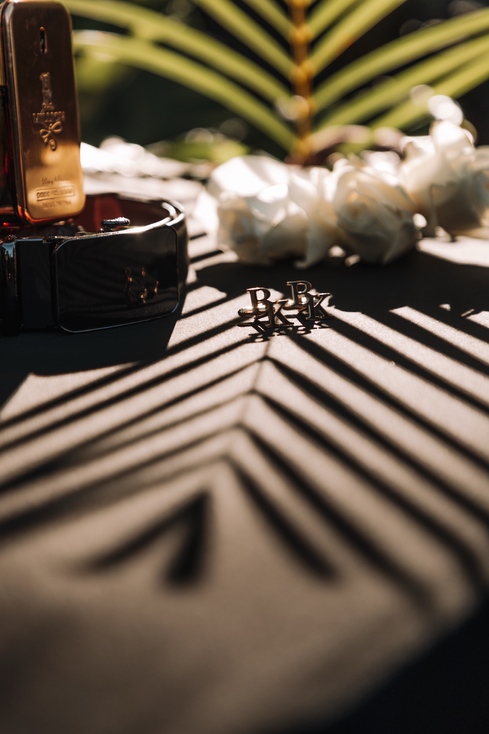 La photo montre un collier en or avec une croix, des fleurs blanches et des initiales « BK » posées sur une surface sombre, éclairées par la lumière n